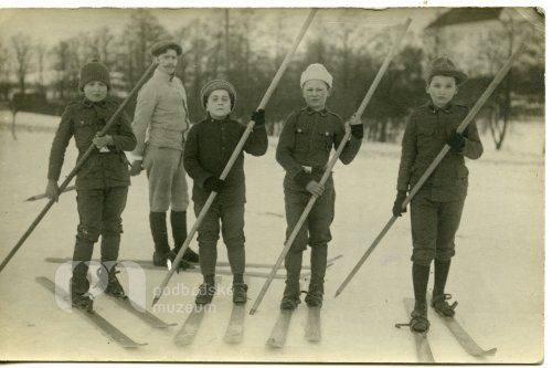 Čtyři chlapci v uniformách na lyžích, v pozadí muž na lyžích a rožmitálský zámek
